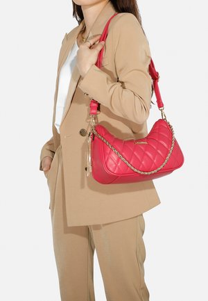 Borsa a tracolla trapuntata rossa con catena e dettaglio a frange, caratterizzata da una forma curva e una texture in pelle liscia, tenuta da una persona in un completo beige.