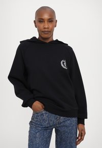 Felpa nera con cappuccio, tasca anteriore, polsini a coste e un piccolo logo bianco sul petto, abbinata a jeans blu brillanti.