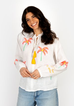 Smiling woman wearing white blouse with pink and orange embroidered palm trees, yellow tassel ties, and light blue jeans on white background.