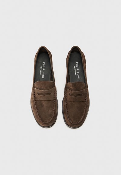 Mocasines de ante marrón con detalles de costura, puntera redondeada y un acabado suave. El interior cuenta con una plantilla acolchada negra.