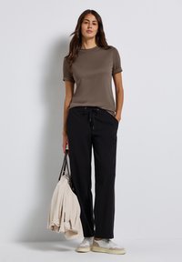 Brun t-shirt med kort ärm, svarta byxor med dragsko, vita sneakers, hållande en ljusfärgad tröja och en väska. Enkel, avslappnad outfit.
