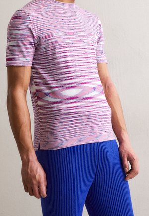 Man draagt een roze-wit patroon overhemd met korte mouwen en gestructureerde felblauwe broek, staand tegen een effen achtergrond.