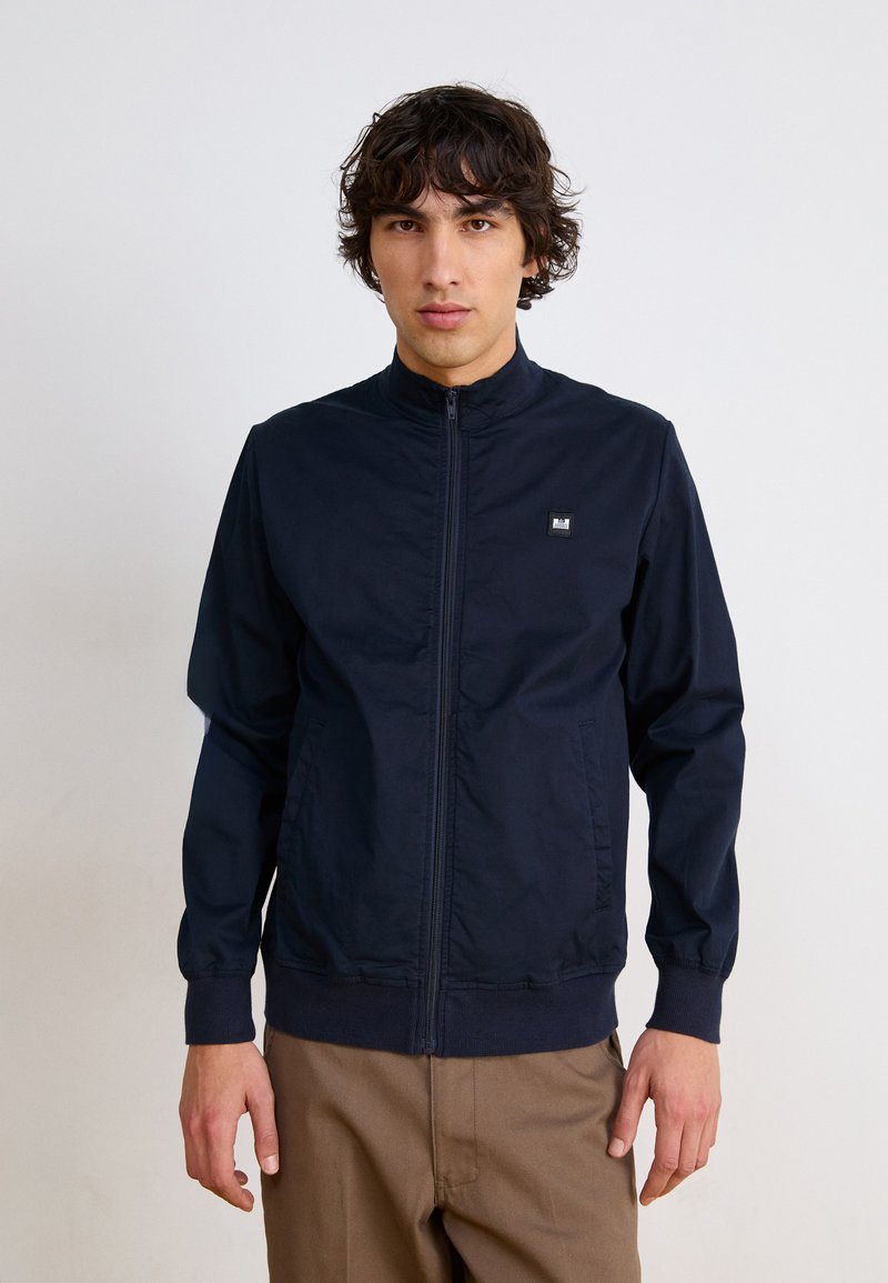 Marineblå zip-up jakke med høy krage, elastiske mansjetter og hem, med en liten logo på venstre bryst. Glatt, lett materiale.