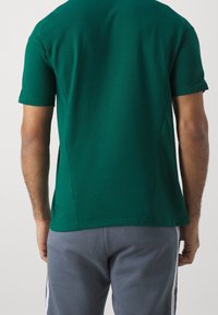 Zelené tričko s krátkým rukávem a kulatým výstřihem, s hladkou texturou a bočními rozparky, spárované se šedými kalhotami s bílými pruhy.