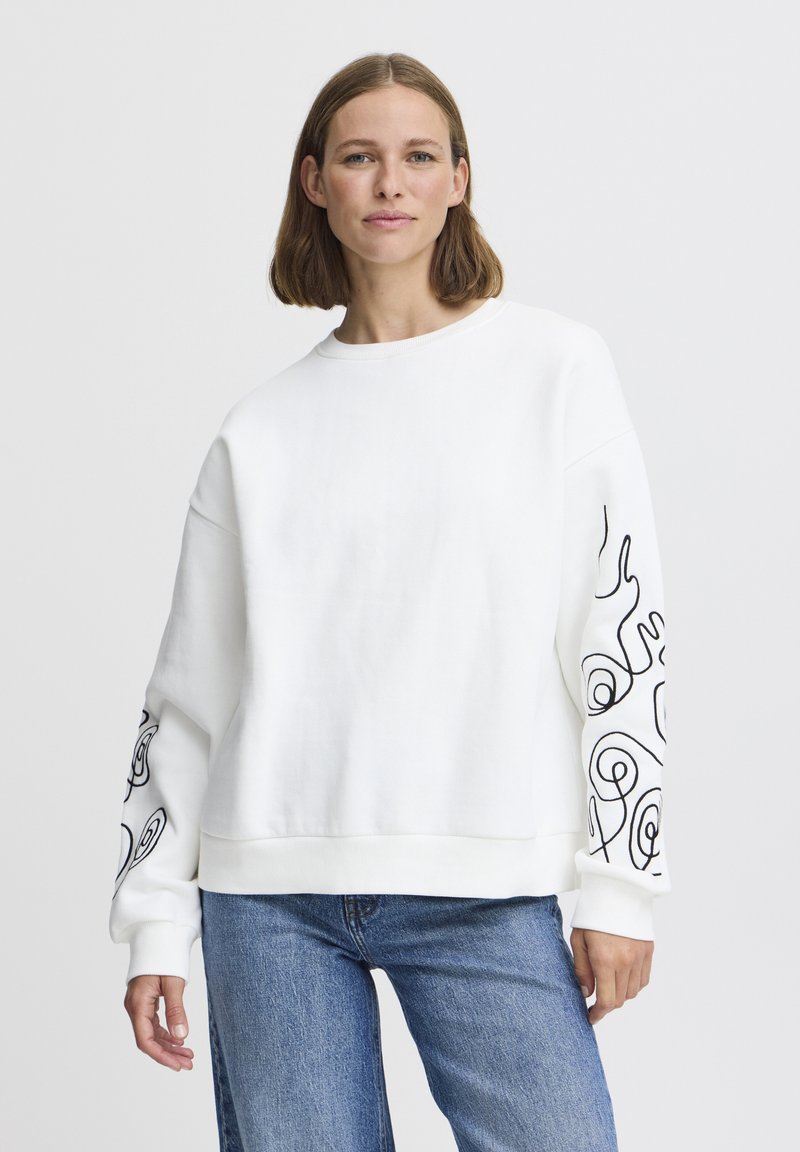 Jeune femme aux cheveux bruns mi-longs portant un sweat blanc avec des motifs abstraits noirs sur les manches et un jean bleu, debout.