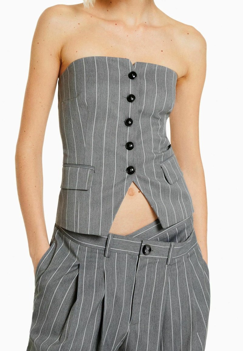 Bezramkowy szary garnitur w paski z czarnymi guzikami, z dopasowaną górą z rozcięciem z przodu i luźnymi, wysokimi spodniami z kieszeniami.