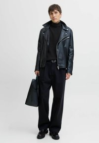 Chaqueta de motorista de cuero negro sobre un cuello de tortuga negro acanalado, combinada con pantalones holgados negros y una bolsa tote negra. Zapatos brillantes completan el look.