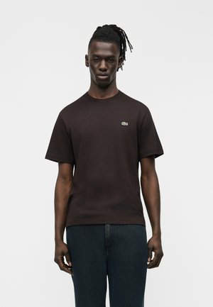 Joven de piel oscura y rastas, vestido con una camiseta negra lisa de manga corta con un pequeño logo de cocodrilo en el pecho, de pie frente a un fondo blanco.