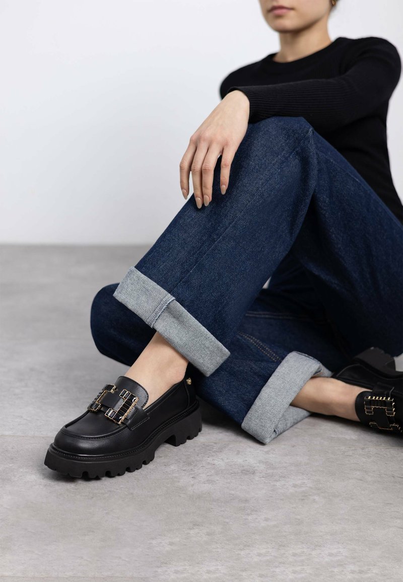 Mocasines de cuero negro con herrajes dorados, suela gruesa y detalles texturizados. Llevados con jeans oscurecidos de mezclilla con dobladillo enrollado. Acabado suave.