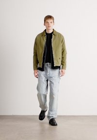Young man walking indoors wearing an olive green jacket, black shirt, light-wash ripped jeans, and black shoes against a plain white wall.
