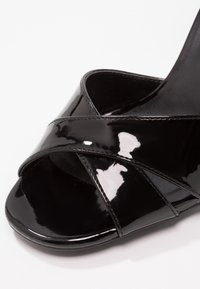 Sandale en cuir verni noir avec larges brides croisées et bout ouvert, présentée sur fond blanc.