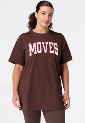 Vrouw met krullend haar draagt een bruin T-shirt met het woord "MOVES" in grote roze letters, staand tegen een effen achtergrond.
