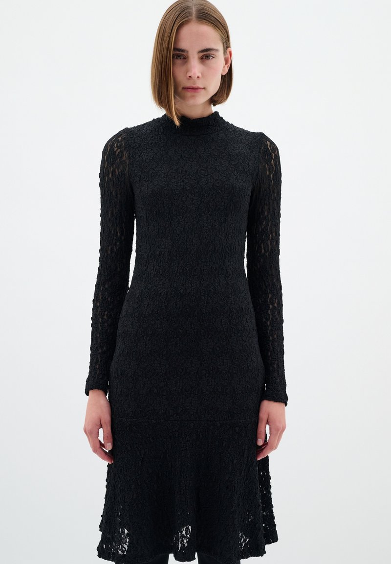 Rochie neagră din dantelă cu mâneci lungi, decolteu înalt și bust ajustat. Prezintă un tiv evazat și un model floral texturat pe întreaga suprafață.