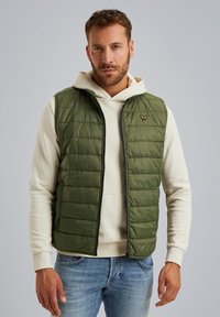 Grön pufferväst med quiltat mönster, kontrasterande svart dragkedja och emblem på bröstet, bärs över en beige hoodie och blå jeans.