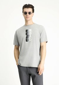 Camiseta de manga corta gris con texto gráfico vertical en negro, gris y blanco. Tejido de algodón suave, corte relajado y cuello redondo.