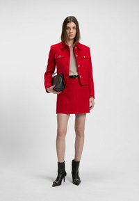 Veste rouge cintrée avec mini-jupe assortie. Haut beige, ceinture noire et petit sac à main noir. Bottines noires à bout pointu avec des accents.