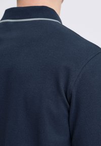 Marinblå pikétröja med textilstruktur, med krage och en kontrasterande ljusgrå rand längs krage kanten.