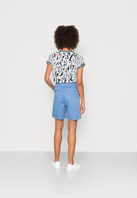 Donna con capelli ricci che indossa una camicia a motivi bianco e nero, pantaloni corti blu e sneakers bianche, in piedi di fronte a un muro bianco.