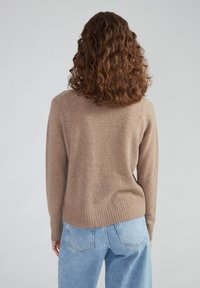 Tan wollen trui met een geribbelde rand, ronde halslijn en een ontspannen pasvorm, gecombineerd met lichtblauwe denim jeans. Gezien vanuit de achterkant.