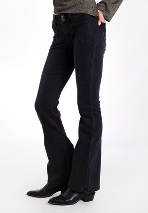 Los jeans acampanados negros tienen una cintura alta con cierre de doble botón, cuatro bolsillos y sutiles detalles de costura. Combinados con botas negras de tacón.