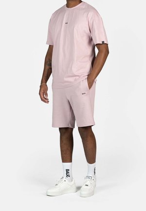 Homme portant une chemise à manches courtes rose clair et un short assorti, avec des chaussettes BLAR blanches et des baskets blanches, se tenant avec une main dans sa poche.