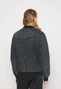 Svart denimjacka med en texturerad yta, utrustad med en rund krage, ribbade ärmslut och en framträdande Nike-logotyp präglad på baksidan.