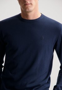 Marineblaue Langarm-T-Shirt mit Rundhalsausschnitt, aus weichem Stoff gefertigt und mit einem dezenten gestickten Logo auf der linken Brust ausgestattet.
