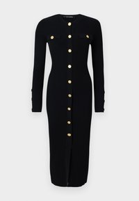 Robe à manches longues en ribbed noir avec des fermetures à boutons dorés, deux poches poitrine et une fente frontale. Texture douce, forme ajustée.