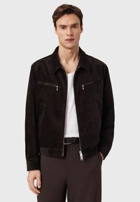 Καφέ σακάκι από σουέντ με φερμουάρ σε τσέπες στο στήθος, επίπεδη γιακά και πλαϊνές τσέπες, συνδυασμένο με λευκό πουκάμισο και σκούρο παντελόνι.