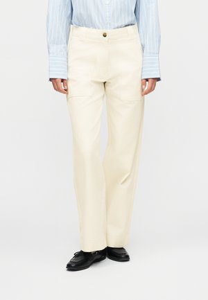 Άτομο που φοράει κρεμ παντελόνι με ίσια μπάτζα και εμπρόσθιες τσέπες με πατζάκια, μπλε και λευκό ριγέ πουκάμισο με μακριά μανίκια και μαύρα μοκασίνια.