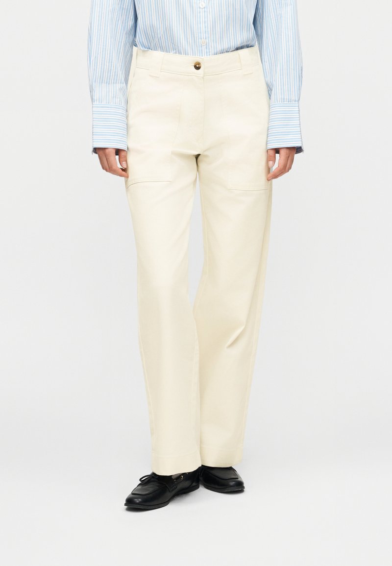 Άτομο που φοράει κρεμ παντελόνι με ίσια μπάτζα και εμπρόσθιες τσέπες με πατζάκια, μπλε και λευκό ριγέ πουκάμισο με μακριά μανίκια και μαύρα μοκασίνια.