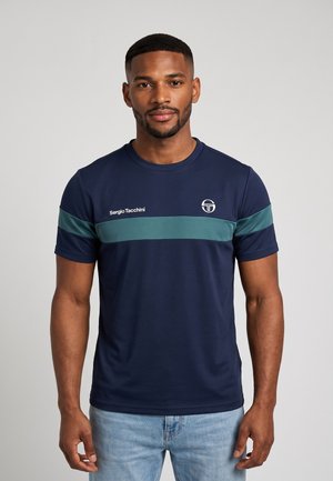 Giovane uomo che indossa una maglietta blu navy Sergio Tacchini con una striscia orizzontale turchese sul petto, in piedi davanti a uno sfondo chiaro e uniforme.