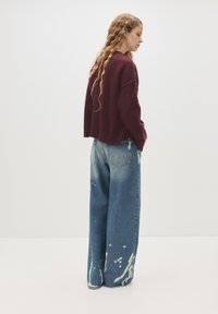 Maglione a maglia bordeaux con vestibilità rilassata. Jeans in denim a vita alta e gamba larga con un motivo tie-dye azzurro chiaro sulle gambe.
