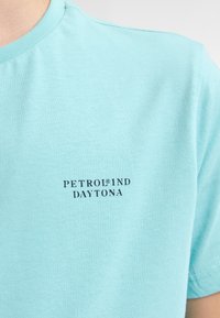 Camiseta de algodón azul claro con cuello redondo y un pequeño texto de logo azul oscuro "PETROLIND DAYTONA" en el pecho. Textura suave.