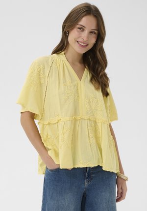 Kobieta z długimi brązowymi włosami w żółtej, haftowanej bluzce z krótkim rękawem i niebieskich dżinsach, uśmiechająca się, z jedną ręką w kieszeni.