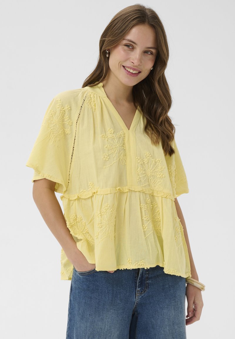 Femme aux longs cheveux bruns portant un chemisier jaune à manches courtes brodé et un jean bleu, souriante, une main dans la poche.