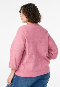 Rožinis ribinis megztinis su laisvo silueto, plačiomis rankovėmis ir apvalia iškirpte. Dėvimas su mėlynais džinsais. Matomas tekstūruotas mezginio raštas.