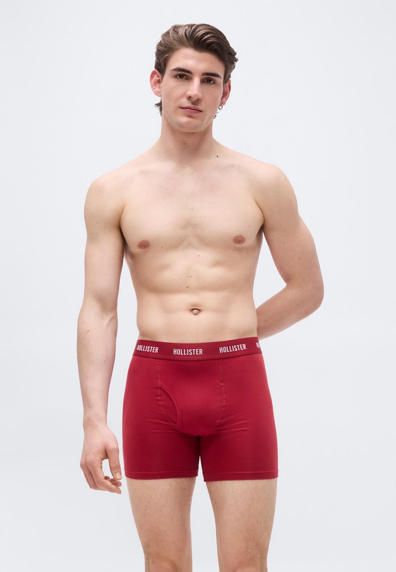 Άντρας χωρίς πουκάμισο με κοντά καστανά μαλλιά, φορώντας κόκκινα εσώρουχα Hollister, στέκεται μπροστά σε απλό φωτεινό φόντο.