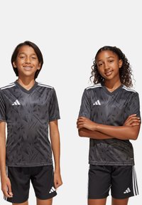Graue Sporttrikots mit angularm Muster, schwarze Shorts und weiße Streifen auf den Schultern. Der Stoff wirkt leicht und atmungsaktiv.