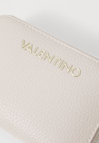 Gaiši bēšs ādas maks ar teksturētu apdari. Izceļas ar zeltītu iepresētu "VALENTINO" logo uz priekšpuses, ar taisnstūra formu.
