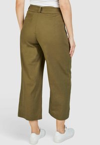 Olivgrüne Leinen-Culotte mit einem mittelhohen Bund, Seitentaschen und einem lockeren Schnitt, kombiniert mit weißen Sportschuhen.