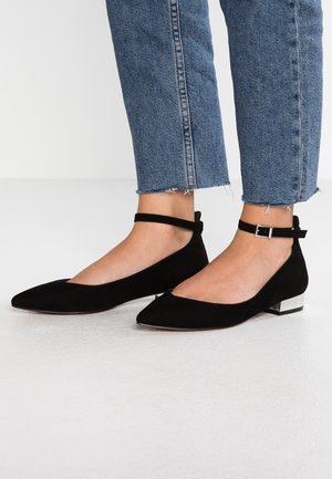 Schwarze Wildleder-Ballerinas mit spitzen Zehen, Knöchelriemen und einem kleinen, metallischen Akzent an der Ferse. Getragen zu gekürzten blauen Jeans.