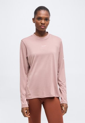 Vrouw draagt een lichtroze shirt met lange mouwen en bruine legging en staat tegen een effen witte achtergrond.