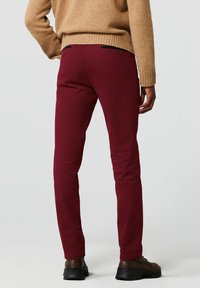 Bordeauxrode broek met een slanke pasvorm, voorzien van achterzakken met zwarte accenten en een gladde stoftextuur, gecombineerd met donker schoeisel.