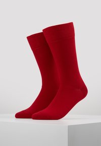 Rote Socken mit einem gerippten Bündchen, glatter Textur und einer standardmäßigen Crew-Länge, aufrecht stehend auf einem weißen Sockel vor einem neutralen Hintergrund.
