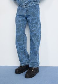 Blå denimjeans med et tonalt blomsterdesign, lige skæring, kombineret med sorte sko med gyldne snørehuller og en tekstureret sål.