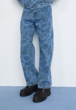 Blå denimjeans med et tonalt blomsterdesign, lige skæring, kombineret med sorte sko med gyldne snørehuller og en tekstureret sål.