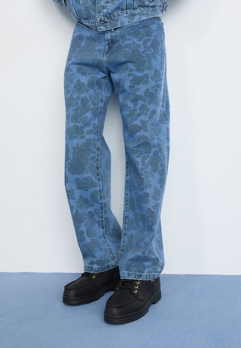 Blå denimjeans med et tonalt blomsterdesign, lige skæring, kombineret med sorte sko med gyldne snørehuller og en tekstureret sål.
