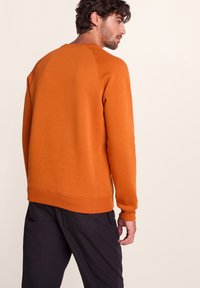 Man with dark hair wearing an orange long-sleeve sweatshirt and black pants, looking to the side against a plain light background.