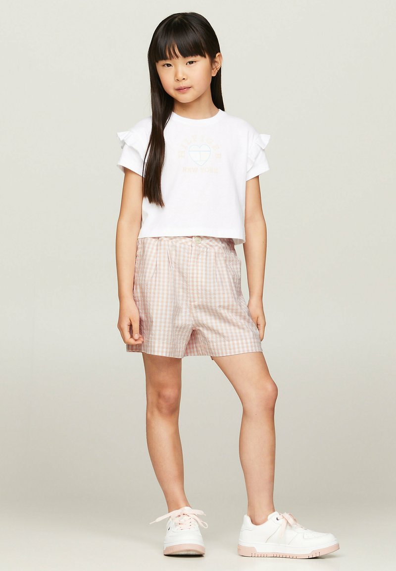 Weißes Baumwoll-T-Shirt mit Rüscheärmeln und pastellfarbenen Grafiken, kombiniert mit rosa karierten Shorts mit Knopf und flachem Vorderteil. Weiße Sneakers.